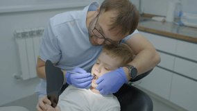 Professional dentist man with glasses shows the boy in mirror baby teeth and gums. Boy is sitting in dental chair and carefully looks in mirror. Slow motion