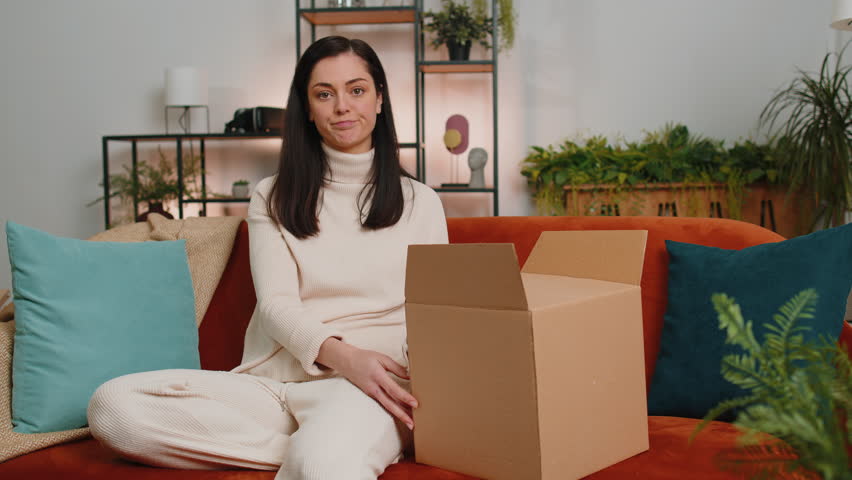 Angry dissatisfied shopper young woman unpacking parcel feeling upset and confused with the wrong delivery from an online store, bad quality purchase at home. Girl indoors in living room sits on couch | Shutterstock HD Video #1099254097