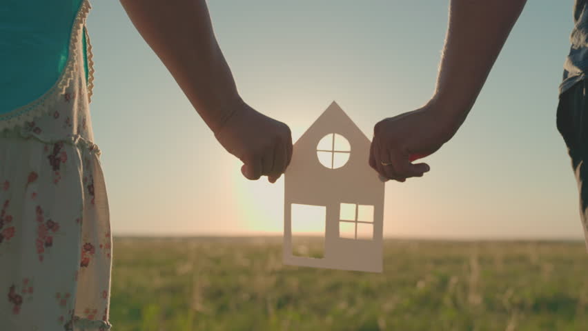 The family's hands are holding a paper house at sunset, the sun is shining through the window. The symbol of the house, happiness. The concept of building a house for the family. Dream to buy a house Royalty-Free Stock Footage #1099254477