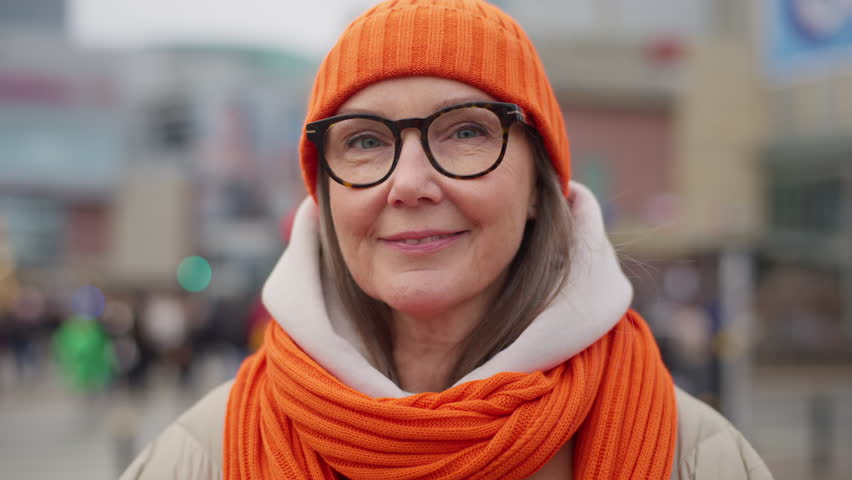 Portrait urban style happy middle aged older lady standing on city street. Beautiful senior woman in trendy winter outfit wearing bright orange hat and scarf backdrop people crowd crossing crosswalk