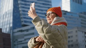 Happy mature female takes photo or video using smartphone camera standing in the middle of downtown urban city landscape. Senior woman, blogging, street photography, modern architecture