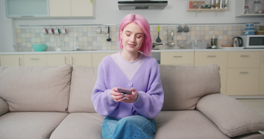 Teenager girl blogger sits on cozy sofa and scrolls social media on smartphone. Pink-haired blogger follows events and newness on Internet | Shutterstock HD Video #1099261315