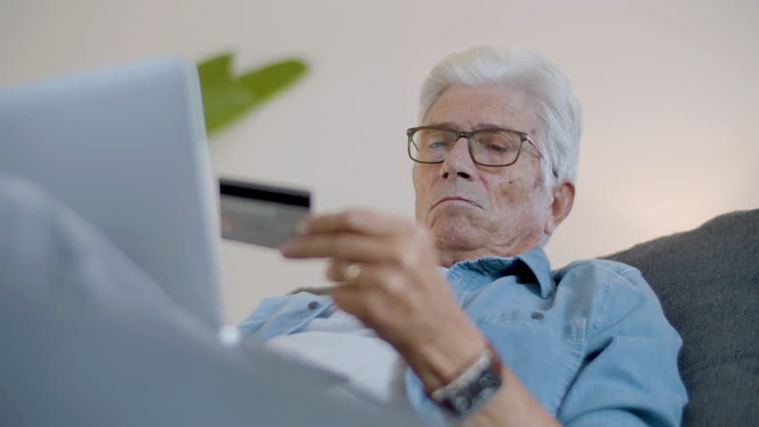 Senior man entering credit card details to do online shopping. Medium shot of focused mature man making purchases through internet using laptop while lying on sofa. Digital device, e-commerce concept | Shutterstock HD Video #1099269203