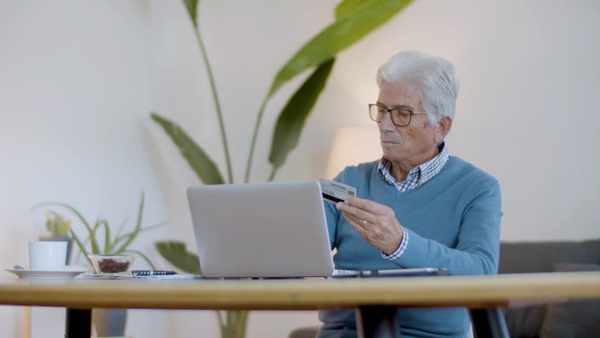 Senior man making purchases online, paying with credit card. Focused Caucasian pensioner sitting at table, entering debit card details website of online store. E-commerce, digital devices concept | Shutterstock HD Video #1099269237