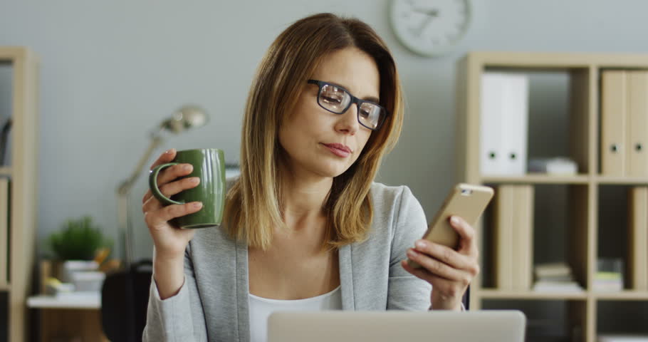 Close up of the Caucasian businesswoman in glasses scrolling on her smartphone while drinking coffee in the office during lunch break. | Shutterstock HD Video #1099269919