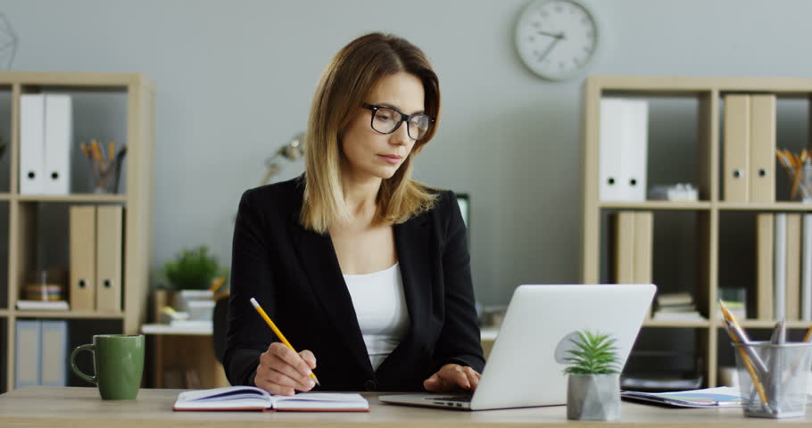 Beautiful female office worker in the glasses working at the laptop computer and writing some data on the paper sheet in the nice modern office.