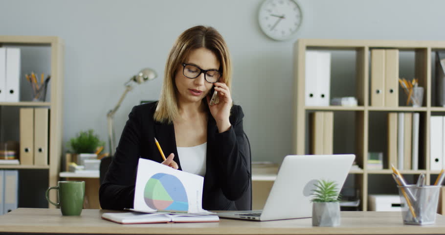Caucasian pretty woman in glasses sitting at the desc with a laptop and working with documents while speaking on the phone in the office. | Shutterstock HD Video #1099269933