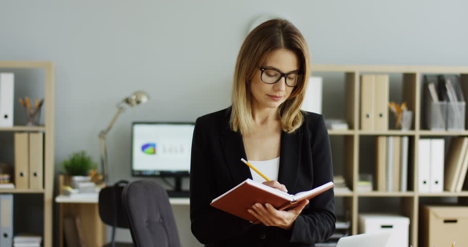 Portrait shot of the young pretty woman with planner notebook in hands standing and smiling to the camera in the modern office room. Inside