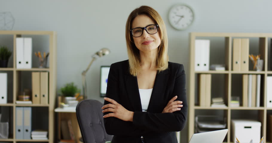 Caucasian good looking businesswoman in the black jacket and glasses standingin her office room and crossing her hands in front her while smailing to the camera.
