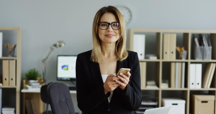 Attractive young woman in the glasses standing in the office at her workplace and scrolling on the smartphone, then looking at the camera.