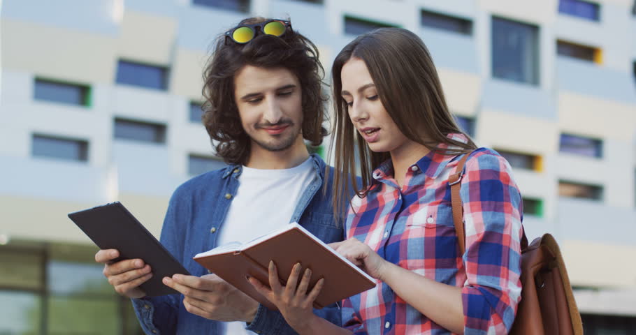 Portrait of the young caucasian couple talking near their college and looking at the book or notebook and tablet computer in hands. Outside.