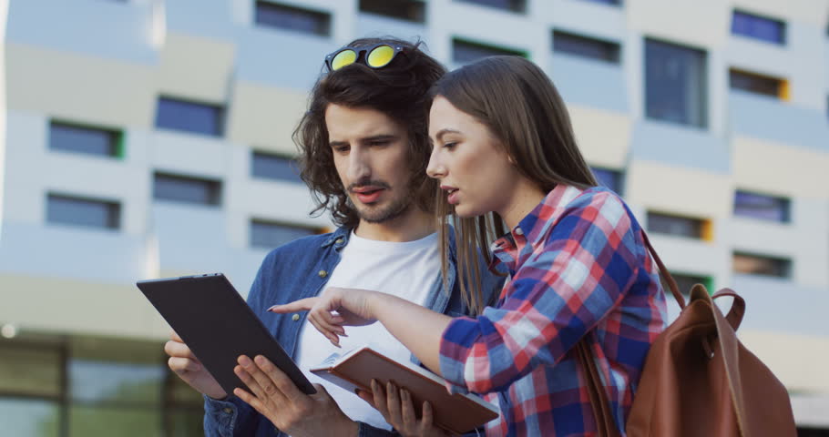 Close up of the couple of the male and female students standing together near their college and looking for something in the tablet device and book in hands. Outdoors.
