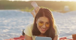 Portrait of the beautiful caucasian young woman in the sunglasses smiling while watching something on the tablet computer at the shore. Outdoors.