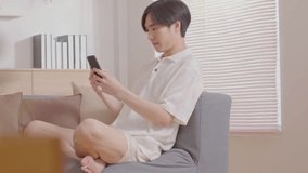Handsome Freelancer Asian young man sit on couch looking on mobile screen recieved good news raise hand up celabrating. Happiness using mobile phone surfing internet or playing game at home