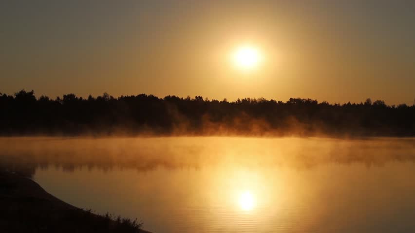 Sunrise above the lake. Sandy beach. Morning fog, mist over the water. Early summer morning.