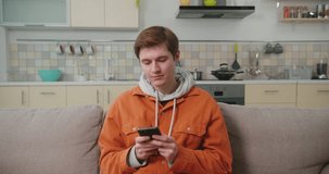Young man scrolls social media on smartphone sitting on cozy sofa. Red-haired blogger enjoys spending time exploring social networks on Internet