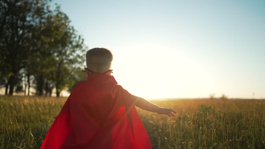 Boy in superhero costume. Child in red raincoat and mask runs through grass in park. Kid plays superhero in park at sunset. Child in suit runs through green grass at sunset. summer outdoor recreation | Shutterstock HD Video #1099280389