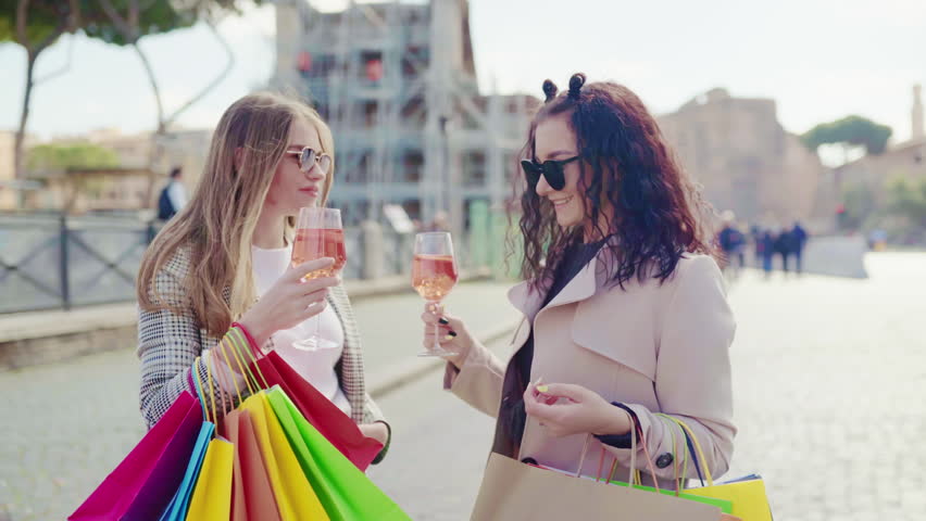 Female friends meeting in the city centre and drinking refreshing aperol spritz cocktail after perfect shopping day, smiling ladies talking and flirting on camera, stylish women with colourful