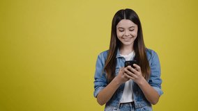 Cute Caucasian teen girl holding smartphone and smiling at camera while pointing with her index finger at copy advertising space for your promotional text or video, over a yellow background. Mockup.