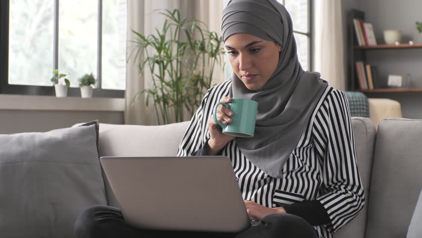 Focused muslim female at computer sits on couch at home,thoughtful pensive young woman working at the laptop sitting on sofa,hard decision problem solving | Shutterstock HD Video #1099285811