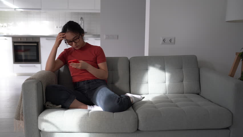 Upset disappointed Asian woman sitting on sofa, holding cellphone in hands, getting angry and frustrated with poor internet connection on mobile phone, receiving bad news, annoyed by broken gadget | Shutterstock HD Video #1099288229