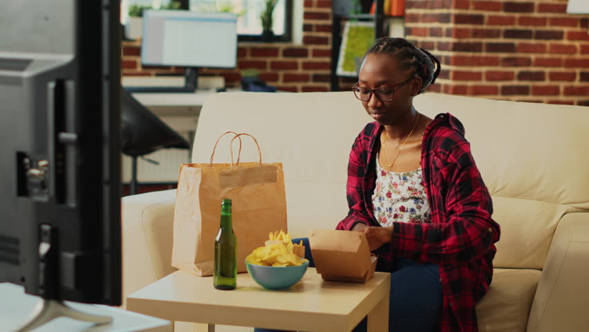 Happy woman eating hamburgers with fries and beer, having fun with action film on television. Smiling person enjoying burger and snacks in bowl, binge watching tv show in living room.