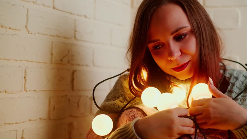 Young sad woman in knitted sweater wrapped in garland of glowing bulbs sitting on floor in room. Unhappy female looking down, sitting, swaying in dark with light on bulbs turned on.