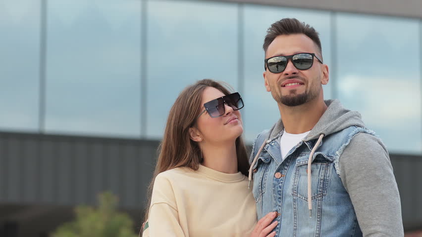 Front view of boy and girl wearing sunglasses, having rest at resort. Young, happy couple standing, hugging each other, admiring landscape. Concept of leisure and recreation.