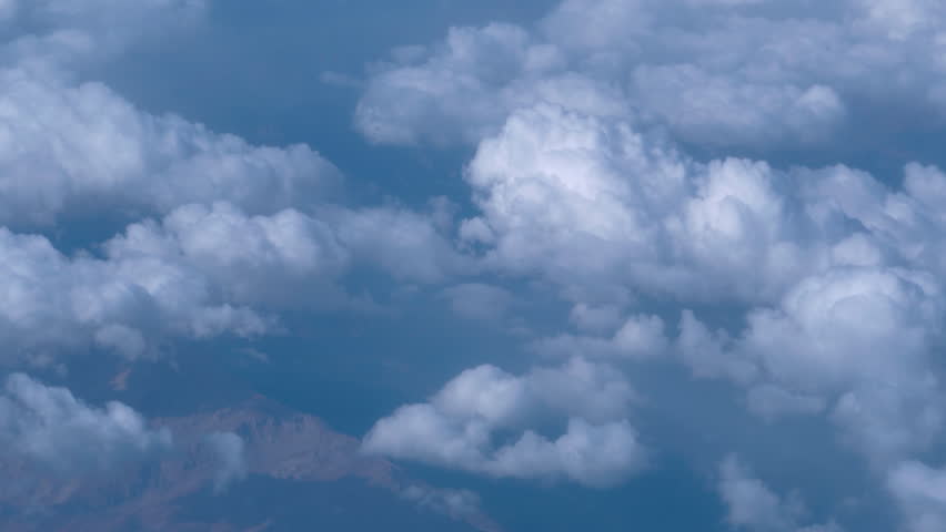 Travel flying above bed of puffy clouds, looking to the left of the frame. Passenger window view aboard an airplane cruising at high altitude.