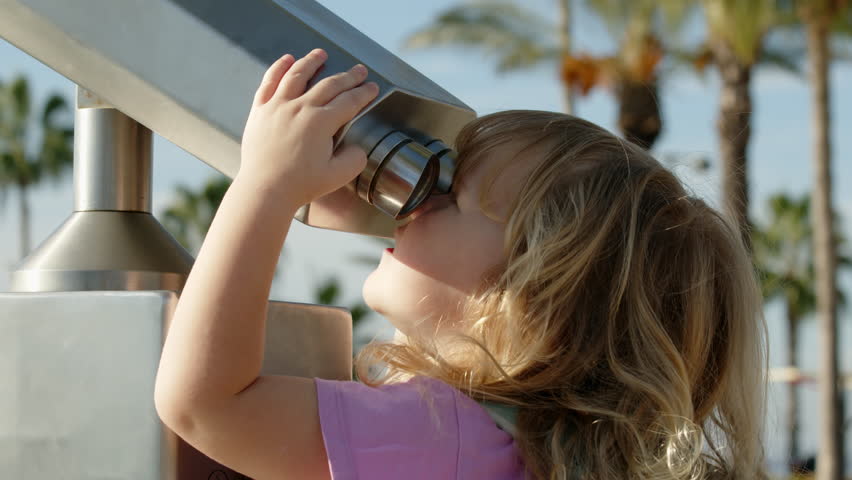 child in pink T-shirt looks through stationary city binoculars in park. Funny curly blonde girl holds her hands on large binoculars after paying with coins. Beautiful view through spyglass in city.