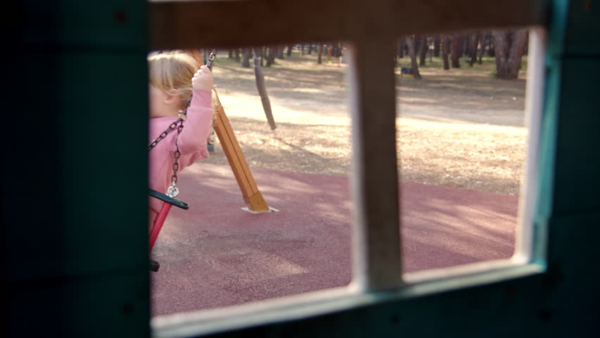 Child in pink suit swings on swing in playground. View from children house window to girl who is having fun. | Shutterstock HD Video #1099306001