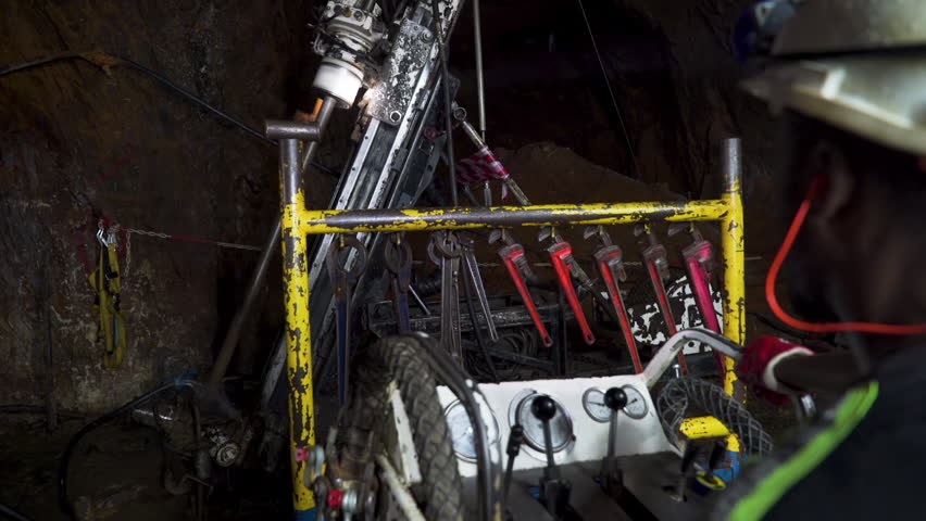 An African miner excavates rock operating an underground drill rig in a deep ore mine.