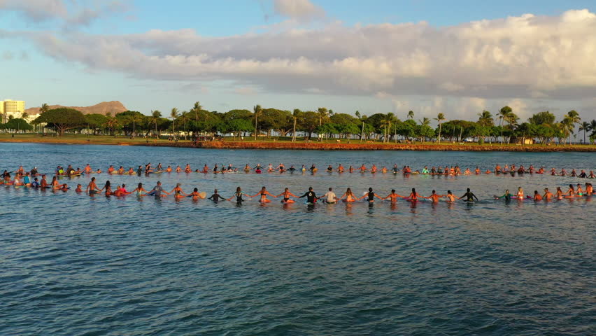 Panning Shot Of Surfers On Surfboards Holding Hands In Sea By Buildings Under Clouds - Oahu, Hawaii