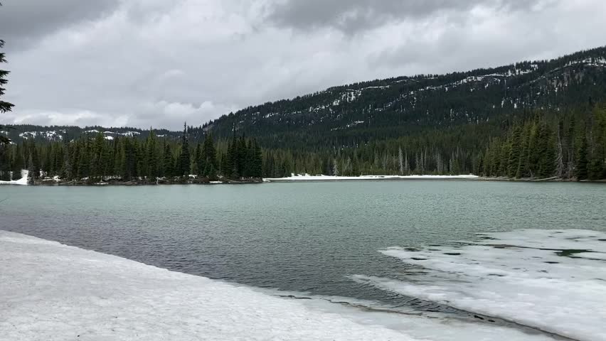View of partially frozen Devils Lake with forest of evergreens in the background on the Cascade Lakes Scenic Byway. Taken on a cold, cloudy day - Nr Bend, Oregon, USA