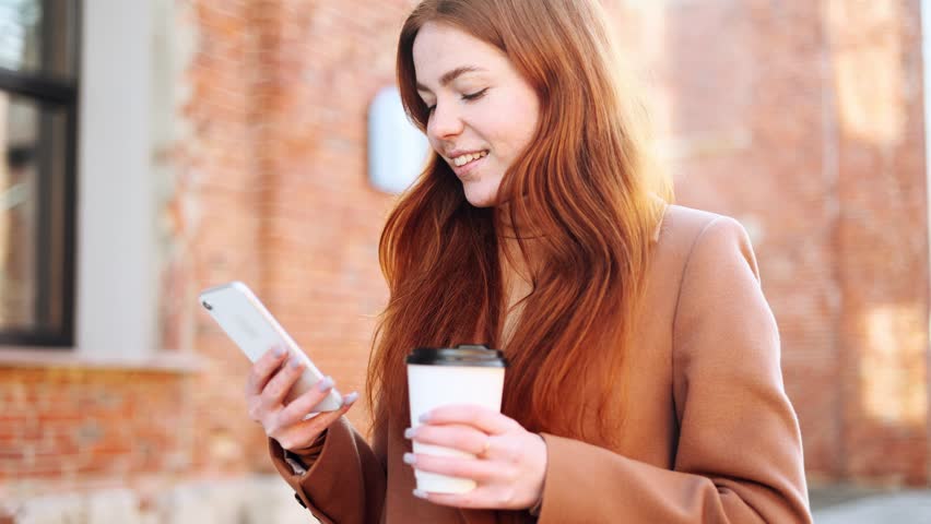 Portrait of smiling red haired ginger young woman with cup of coffee or tea hold smartphone scrolling social media texting browsing online on the street in brick city centre alone | Shutterstock HD Video #1099319065