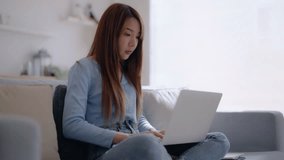 Young asian woman using computer laptop and mobile smartphone while seated on couch at home