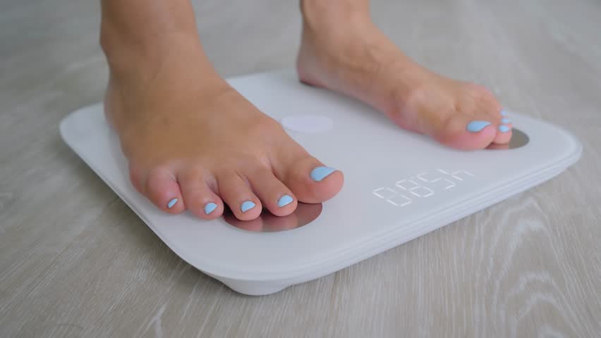 Woman weighing herself - female bare feet stepping on white digital floor scales at home: close up view. Measuring weight, control, wellness and diet concept | Shutterstock HD Video #1099335483