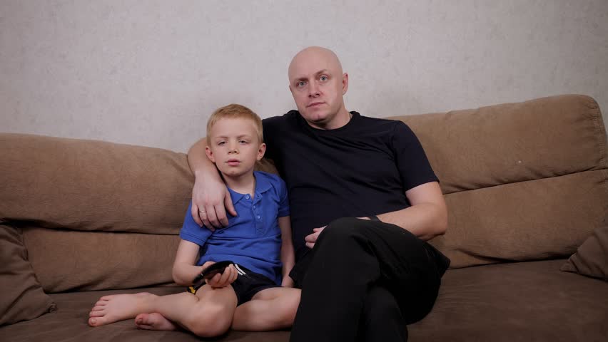 Happy father and son are using the remote control and watching TV while sitting at home on the couch. | Shutterstock HD Video #1099337433