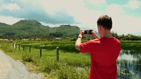 Traveler in Asia captures stunning natural beauty with mobile phone. Back view shows millennial blogger or vlogger in red T-shirt filming green mountains. Perfect vacation footage.