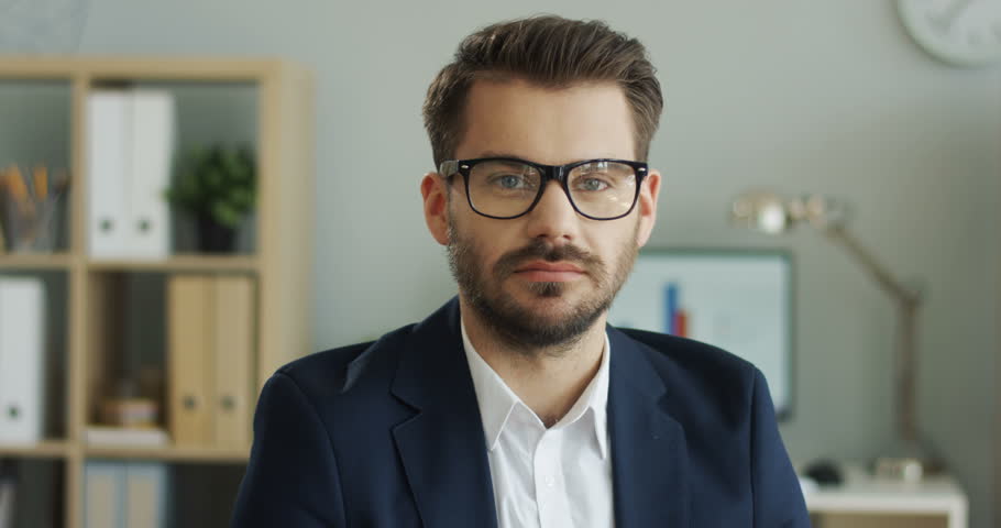 Portrait of the handsome young Caucasian man in glasses, shirt and jacket smiling tothe camera in his office. Close up. | Shutterstock HD Video #1099340427