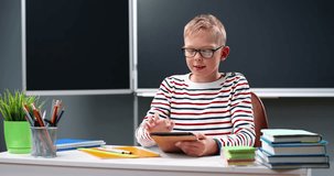 Portrait of Caucasian boy in glasses sitting at table a school, smiling to camera and using tablet device. Teen schoolboy in eyeglasses taping and watching video on gadget. Teenage playing on computer