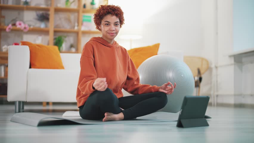Portrait of smiling young adult american woman sitting on mat for sports in yoga meditation pose looks at camera. Cheerful female relaxing at home. Healthy lifestyle and domestic leisure activity. | Shutterstock HD Video #1099341203