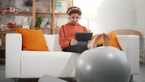 Young attractive african american woman in sportswear sit at couch watching fitness online video on digital tablet and put her feet on fitness ball in living room. Home workout and fitness exercise.