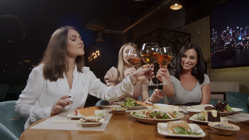 Three cute girlfriends having a rest sitting at a table and drinking wine merrily talking. Girls clink glasses with wine glasses.
 | Shutterstock HD Video #1099343015