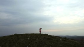 Drone footage of a 30s one woman with red tights and tops dancing on a Tumulus hill on a cloudy sky background