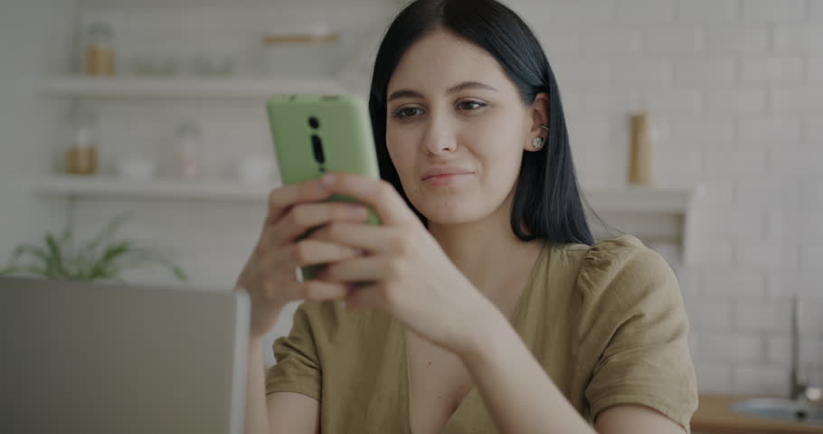 Portrait of cheerful young lady using smartphone smiling enjoying social media in kitchen at home. Modern technology and communication concept. | Shutterstock HD Video #1099344885