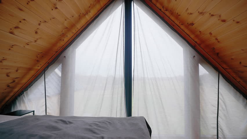 A woman dressed in gray enters an attic room. The ceiling is wooden. The woman opens the curtains. High quality 4k footage