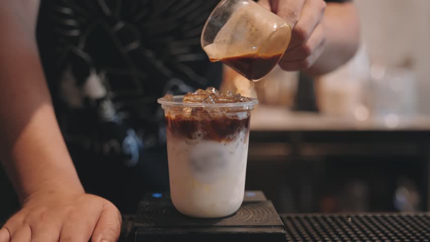 Barista pouring latte dark coffee with milk in plastic glass on counter bar at coffee shop | Shutterstock HD Video #1099345647