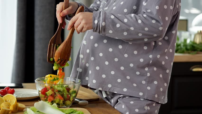 Pregnant housewife wearing pyjama prepares yummy dish at home. Woman uses wooden spoons and mixes chopped organic vegetables in healthy salad, slow motion | Shutterstock HD Video #1099346749