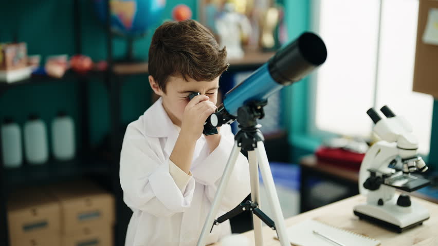 Adorable hispanic boy student using telescope writing on notebook at laboratory classroom